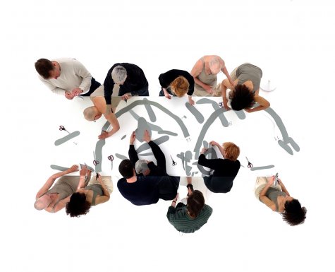 Eine Gruppe von Menschen sitzt oder steht um einen großen Tisch und arbeitet gemeinsam an einer weißen Fläche mit aufgedruckten grauen Linien und Zahlen. Sie benutzen Scheren, um Teile entlang der Linien auszuschneiden. Die Szene wird von oben aufgenommen.