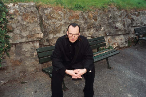 Das Porträt zeigt einen schwarz gekleideten Mann, der breitbeinig auf einer Parkbank mit gefalteten Händen sitzt. Er lehnt sich nach vorne, trägt eine Brille und schaut den Betrachter direkt an.