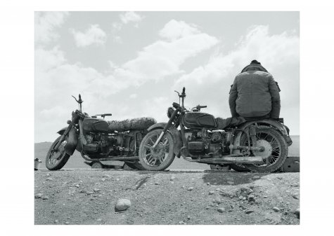 Die Schwarzweißfotografie zeigt zwei Fahrräder am Rand einer Straße, die durch die Wüste führt. Auf dem rechten Fahrrad sitzt ein Mann in Winterkleidung mit dem Rücken zum Betrachter.