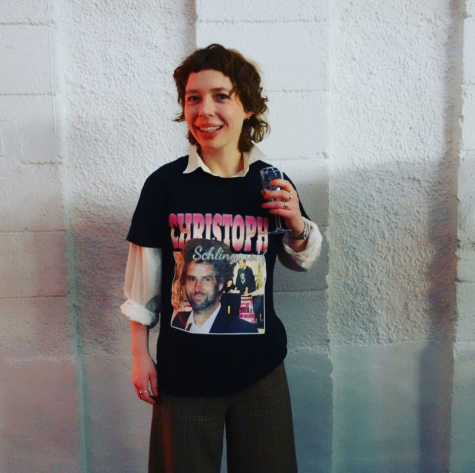 Das Porträt zeigt eine junge Frau im fast vollständigen Profil vor weißem Hintergrund. Sie grinst in die Kamera und hält ein Sektglas in der rechten Hand. Auf ihrem schwarzen T-Shirt steht „Christoph“.