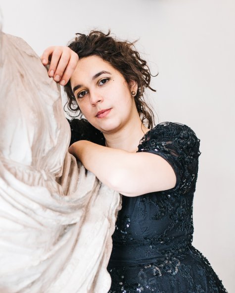 Die Porträtfotografie zeigt eine Frau im schwarzen Kleid, die sich auf eine Skulptur abstützt. Sie hat braune, zurückgesteckte Haare und braune Augen und lächelt leicht in die Kamera.