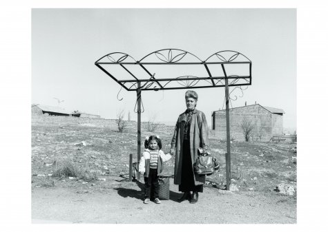 Die Schwarzweißfotografie zeigt eine Frau, die ein Kind an der Hand hält. Sie stehen vor einer zerstörten Bushaltestelle und einer verlassenen Landschaft.