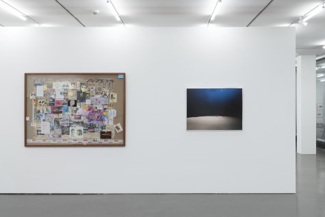 Die Ausstellungsansicht zeigt zwei großformatige Bilder, die aus frontaler Perspektive eingerahmt und nebeneinander aufgehängt sind. Das linke Bild ist eine Collage aus Fotografien, Zeichnungen und anderen Fragmenten, die gemeinsam auf einer Karte angeordnet sind. Das rechte Bild ist eine Fotografie einer Landschaft, die wie eine Wüste wirkt. Das Licht beleuchtet dabei nur den Vordergrund und zeichnet eine nahe Horizontlinie nach. Die Bildmitte ist tiefschwarz und verläuft nach oben hin in ein dunkles Blau.