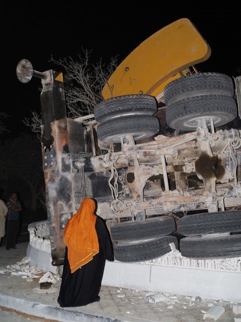 Die Fotografie zeigt eine Frau in schwarzer Kleidung mit orangefarbener Burka, von hinten gesehen. Sie steht vor einem umgestürzten Lastwagen, der aus der Dunkelheit herausragt.