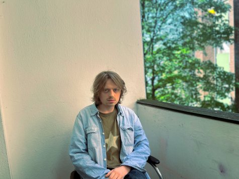 Die Fotografie zeigt einen Mann mittleren Alters, der auf einem Stuhl auf einem Balkon sitzt. Er blickt direkt in die Kamera, mit neutralem Gesichtsausdruck.