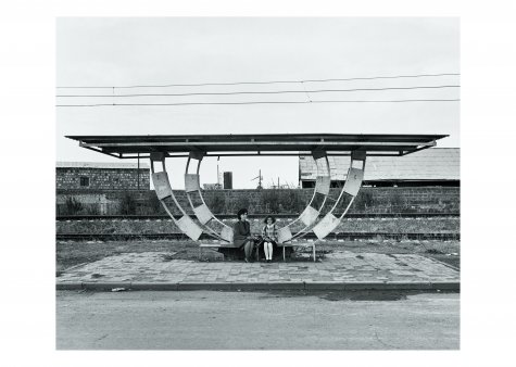 Die Schwarzweißfotografie zeigt zwei Personen, die an einer Bushaltestelle sitzen und in die Kamera blicken. Im Hintergrund sind verlassene Bahngleise zu sehen.