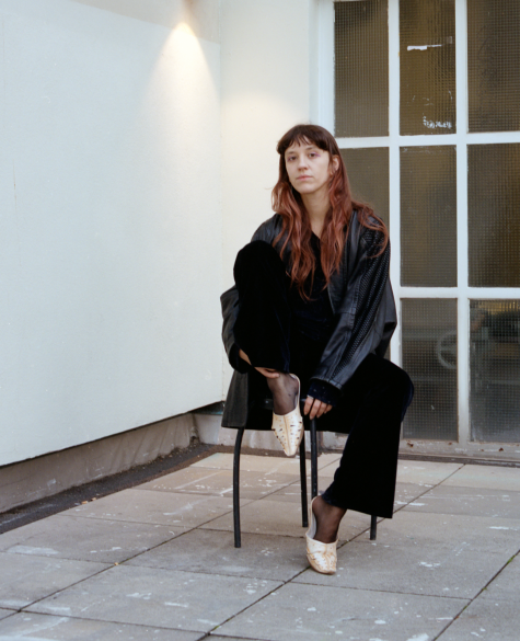 Fotografie einer brünetten, langhaaringen weißen Frau, die, ein Bein angezogen und ernst blickend, auf einem Hocker sitzt..