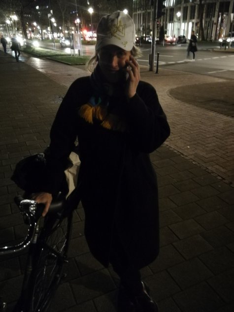 Die Fotografie zeigt eine Frau, die mit der rechten Hand ihr Fahrrad hält und mit der linken ein Handy, scheinbar in ein Gespräch vertieft. Sie blickt lächelnd in die Kamera. Ihr Gesicht liegt teilweise im Schatten der Szene. Es ist Nacht.