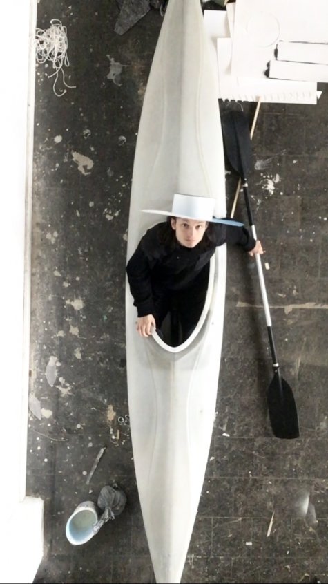 Das Foto wurde aus der Vogelperspektive aufgenommen und zeigt einen Mann mit weißem Hut in einem weißen Kanu, der schwarz gekleidet ist. Das Kanu steht auf dem Boden. Der Mann schaut direkt in die Kamera.