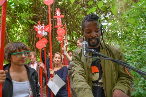 Eine Gruppe von Menschen steht in einem grünen, bewaldeten Außenbereich. Im Vordergrund spricht ein Schwarzer Mann und Dreadlocks in ein Mikrofon. Neben ihm steht ein Mann mit Brille und grünem Gestell. Im Hintergrund halten mehrere Personen rote Stäbe mit runden Schildern und grafischen Symbolen hoch.