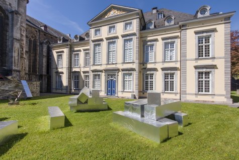 Blick auf die Fassade eines historischen Gebäudes mit symmetrischer Architektur und blauen Doppeltüren; im Vorgarten steht eine Installation aus silbrig glänzenden geometrischen Skulpturen auf dem Rasen.