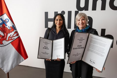Das Foto zeigt Al Qasimi links und Ursula Schulz-Dornburg rechts, die ein aufgeschlagenes Buch präsentieren – den Ernst und Hilla Becher Preis. Sie stehen vor dem Eingang der Ausstellung. Links im Bild ist eine Flagge der Stadt Düsseldorf zu sehen. Beide lächeln in die Kamera.